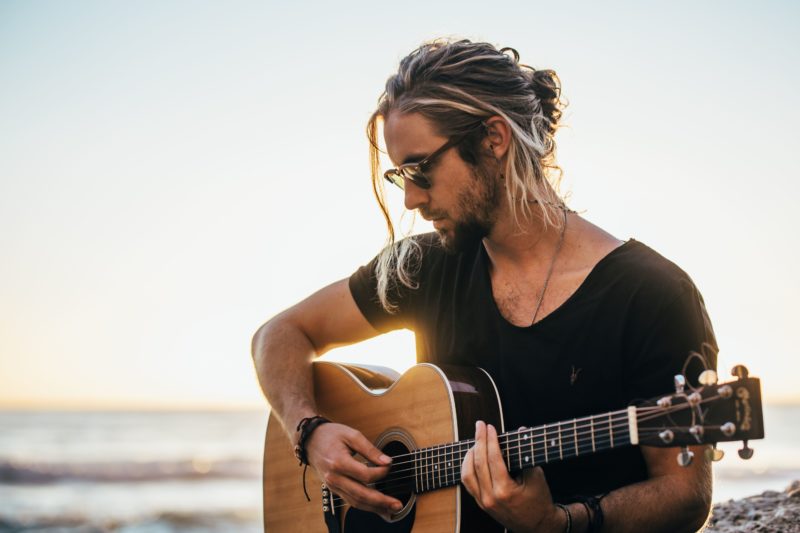 cape town singer Jeremy Loops sitting on a beach playing guitar at sunset with a pair of sunglasses for fans buying jeremy loops tickets