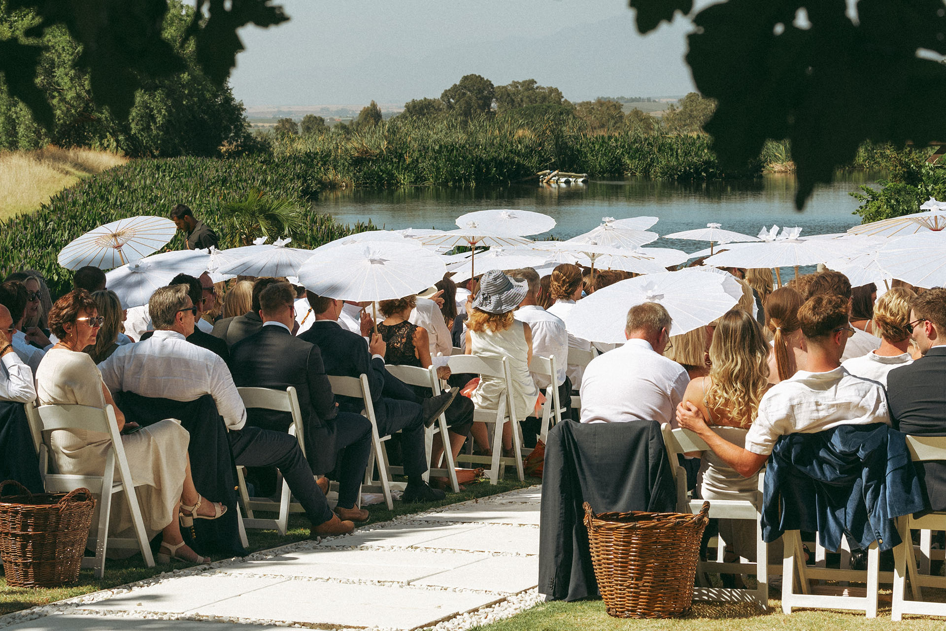 guests sit outdoors under parasols in the summer in the Paarl winelands as they await the bride who chose a wonder summer garden wedding venue at Rhebokskloof.